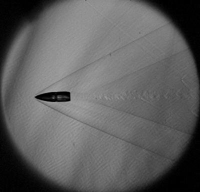 Fotografia schlieren pocisku w locie: wyraźnie widać dziobową falę uderzeniową (bow shock) i fale za pociskiem. Technika schlieren uwidacznia gradienty gęstości, dając intuicyjny obraz frontu fali uderzeniowej. Zdjęcie: Wikimedia Commons