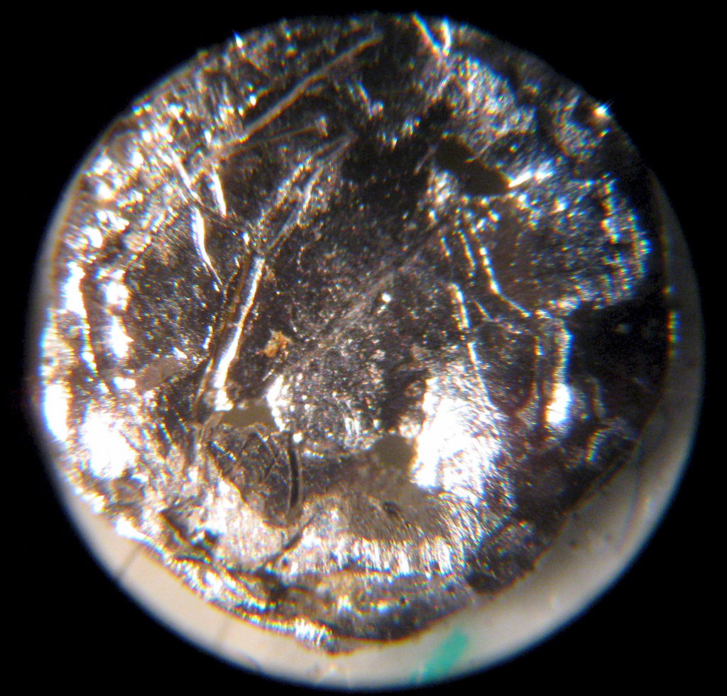 Próbka ameryku-241 widziana pod mikroskopem. Pokazuje cywilne, mierzalne zastosowanie izotopu opisywanego także w kontekście starzenia plutonu. Źródło: Wikimedia Commons, File:Americium_microscope.jpg.